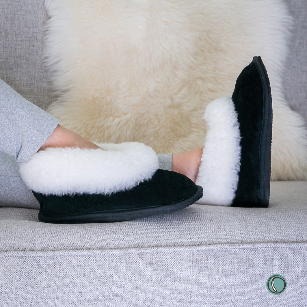 A person wearing black slip-on sheepksin slippers with fluffy white genuine sheepskin, seated on a light grey sofa with a cream sheepskin behind them.